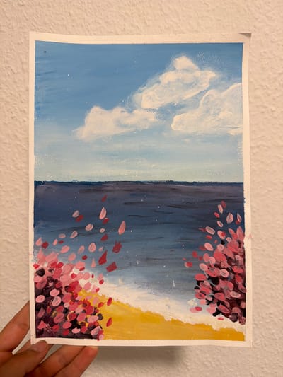 The image is a painting of a beach scene. The sky is a bright blue with fluffy white clouds. The ocean is a deep blue with hints of purple. The beach is a vibrant yellow, with white waves crashing onto the shore. There are clusters of pink and red flowers on the left and right sides of the painting, some petals scattered in the water. The painting is held by a hand with dark skin. The location of this scene is not specified in the image or its context.
