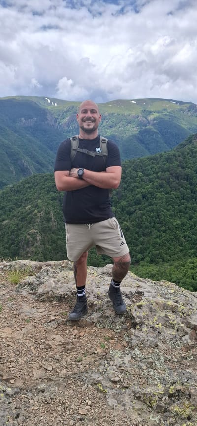A bald, smiling man stands on a rocky outcrop with his arms crossed. He is wearing a black t-shirt, beige shorts, black socks with white stripes, and black hiking boots. He has a backpack on his shoulders and a watch on his left wrist. The background consists of lush green forested mountains under a cloudy sky. The location is identified as a viewpoint in Tazha, Bulgaria.