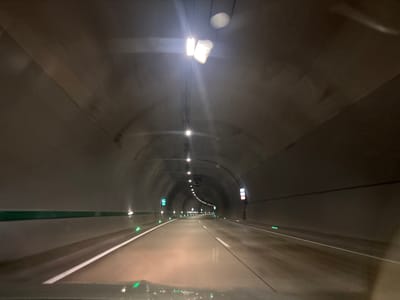 The image shows the interior of a dark tunnel with lights illuminating the road ahead. The tunnel walls are made of concrete and are curved, forming an arched ceiling. There are multiple light fixtures along the ceiling, some of which are bright and cause glare. The road surface appears to be wet and reflective. Green lights are embedded in the road along the sides, marking the edges of the lanes. There are also some signs on the right side of the tunnel. The scene appears to be a view from inside a vehicle moving through the tunnel. There are no people or animals visible in the image. The location is identified as a tunnel in Achara, Georgia.