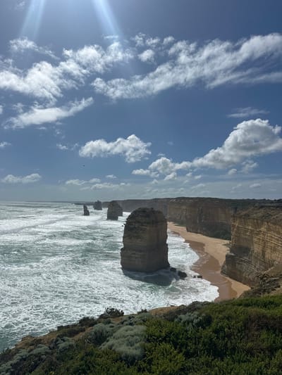 Twelve Apostles rock formations rise from the ocean along a sandy beach bordered by cliffs and green shrubbery. The sea is choppy with white-capped waves. A blue sky filled with scattered white clouds dominates the left side of the image, with the sun shining brightly. The overall scene depicts a coastal landscape under a partly sunny sky.