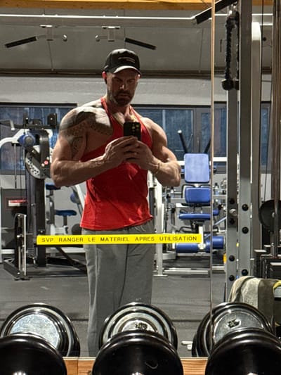 The image depicts an adult male taking a selfie in a gym located in Jemelle, Belgium. The male, appearing muscular, has a beard, wears a black baseball cap, a red tank top, and grey sweatpants. He has a prominent tattoo on his left shoulder and arm. He is holding a dark-colored smartphone horizontally with both hands at chest level, positioned in front of a large mirror, capturing his reflection. A gold-colored chain is visible around his neck, and a gold-colored bracelet is on his right wrist. The background, visible through the mirror's reflection, shows various gym equipment, including cable machines, weight stacks, and benches. In the foreground, a rack of dumbbells is partially visible below the male's waistline. A yellow horizontal sign with black text is reflected across the lower mid-section of the image, reading "SVP RANGER LE MATERIEL APRES UTILISATION," which translates from French as "PLEASE PUT AWAY THE EQUIPMENT AFTER USE." The environment indicates an indoor gym setting with artificial lighting.