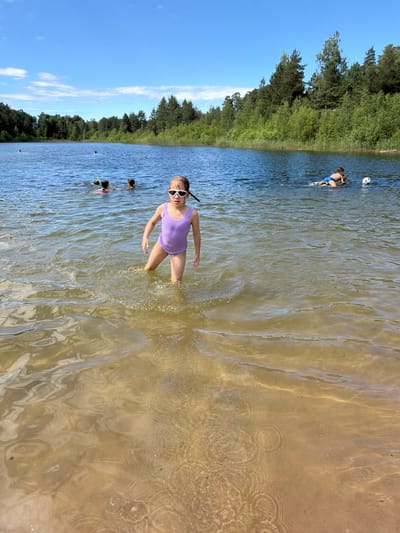 The image depicts a group of people swimming and wading in a lake on a sunny day. In the foreground, a young girl wearing a purple swimsuit and white goggles stands in shallow water, facing the camera. Her body is partially submerged, and ripples emanate from her legs. In the background, several other individuals are visible in the water, some appearing to snorkel. The lake is bordered by a dense line of green trees under a clear blue sky with scattered white clouds. The water is relatively clear, with the sandy bottom visible in some areas, reflecting the sunlight. The overall scene suggests a recreational outing at a natural water body, likely during the summer. The lighting indicates it is daytime, and the clear sky suggests pleasant weather. The specific location context provided is Vētras, Latvia. There is no visible text in the image.