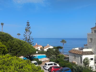 This media file depicts a scenic coastal view in La Gaspara, Spain. The scene is dominated by a bright blue sky and a calm, expansive ocean. In the foreground, lush green trees and shrubs frame the view, with a prominent pine tree reaching upwards. To the right, a white building with a terracotta roof is visible, featuring windows and balconies. A swimming pool with vibrant blue water is situated in the middle ground, surrounded by greenery. Several cars are parked in a lot, including a white van, a red car, and a blue car. In the distance, more buildings with terracotta roofs and palm trees are scattered along the coastline. The sun appears to be shining brightly, suggesting a clear, warm day, likely during daylight hours given the strong illumination. The overall atmosphere is peaceful and evokes a sense of a relaxed vacation setting. No visible text is present in the image.