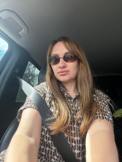 A woman with long, brown hair and sunglasses is sitting in the driver's seat of a car, looking at the camera. She is wearing a brown and white patterned shirt and a seatbelt. The car interior is visible, including the dashboard and a side window showing trees and sky. The scene is well-lit, suggesting it is daytime. The location is Poletaevo 1th, Russia.