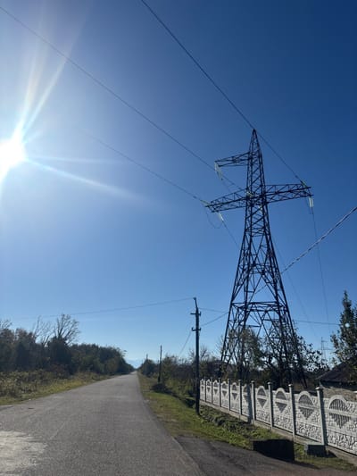 A tall electrical transmission tower dominates the left side of the frame against a clear blue sky with sun glare at the bottom. Power lines stretch from the tower. A paved road runs along the right side with a decorative concrete fence, trees, and other utility poles bordering it. The road recedes into the distance, creating a vanishing point in the background.