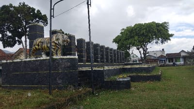 The image captures an outdoor scene dominated by a large tiger statue perched atop a multi-tiered, dark stone structure. The tiger, depicted with white and yellow stripes outlined in black, stands prominently on the highest level of the structure, its head turned slightly to the right with its mouth ajar. The stone structure, possibly a monument or decorative wall, is constructed from dark, rough-hewn blocks and features distinctive yellow horizontal lines painted across its various sections and pillars. These pillars extend in a row across the middle ground.

To the left of the structure, a large evergreen-like tree rises, with glimpses of houses and their red-tiled roofs visible behind it. On the right side, a substantial green tree partially obscures a cluster of buildings, which appear to be residential or small commercial establishments. The foreground and the area to the right are covered in green grass and some weeds. A tall, dark utility pole with overhead wires is visible, extending vertically through the middle-right portion of the frame. The sky is overcast and cloudy, suggesting a daytime scene, possibly with diffused light. The overall setting appears to be a local landmark or a public area within a semi-rural or town environment in Kecamatan Panjalu, Indonesia. There is no visible activity or discernible text.