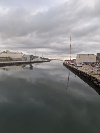 This media file captures an overcast daytime scene of an industrial harbor or canal in Køge, Denmark. The image, correctly oriented, shows a tranquil, grey-green body of water occupying the lower half, with a muted, cloudy sky above.
A long concrete quay or dock stretches from the mid-right foreground into the distance on the left. Along this dock, various industrial structures are visible, including a prominent, large curved concrete silo or building, smaller rectangular sheds, and other operational buildings. Several cars are parked along the quay, suggesting ongoing activity or staff presence. The edge of the dock is lined with dark fendering, possibly old tires.
A tall, light-colored construction crane stands on the dock in the middle ground, its long jib extending towards the left side of the frame. Its reflection is sharply mirrored in the calm water below, alongside reflections of the cloudy sky and distant structures. On the opposite side of the water, further into the background, additional industrial buildings and outlines of distant land can be seen.
The weather is heavily overcast, with a soft, diffused light, indicating a cloudy day without direct sunlight. There are no visible people or discernible specific events taking place; the scene portrays the quiet, functional aspect of a working port. No explicit text is visible. The overall mood is subdued and functional, characteristic of an industrial waterfront on a quiet, cloudy day.
