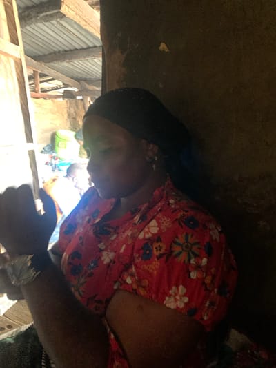 An African woman wearing a black headscarf and a red floral-patterned shirt is shown in profile, looking down and to the left. She has dark skin and appears to be in an indoor setting with natural light streaming in from the left. Behind her, another person in a white shirt is visible, along with some colorful plastic containers. The background appears to be a rustic structure with a corrugated metal roof and wooden beams.