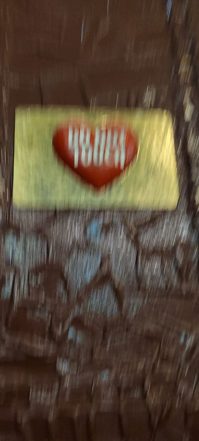 A blurred image shows a red heart-shaped object with white writing on a gold background. The heart is placed on a textured, dark brown surface, possibly chocolate. The text on the heart is difficult to decipher due to the blur, but appears to be "48000". The overall impression is of a decorative item or a wrapper. The location is not discernable from the image itself.