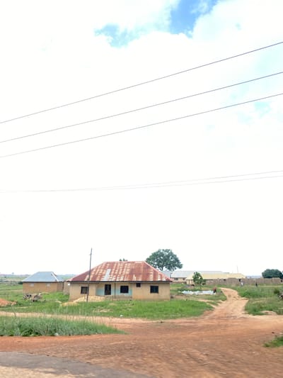 This media file depicts a rural scene in Heipang, Nigeria. The image is oriented vertically, showing a dirt road to the right, with green grass and scattered buildings to the left. In the foreground, a building with a rusty, corrugated metal roof is visible. Further back, other structures with similar roofing materials and some with tin roofs are present, along with stacks of bricks. There are a few people visible in the distance, near the buildings, and some appear to be engaged in activities. Several utility wires stretch across the sky on the left side of the image, which also features a patch of blue sky with some clouds. The overall impression is of a sunny day in a developing community, likely in the afternoon given the light. There is no visible text in the image.