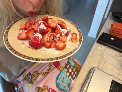 The image shows a close-up view of a person about to eat a plate of sliced strawberries dusted with powdered sugar. The plate has a decorative rim with an orange and brown pattern. The person's mouth is visible, pursed as if about to take a bite. They are wearing a light-colored garment and a pink fabric with a colorful animal print pattern. In the background, to the right, a laptop is partially visible on a countertop. Also on the countertop are two orange boxes labeled "MELLER" and a black wallet. A pair of white earbuds with a tangled cord lies next to the laptop. The environment appears to be indoors, likely a kitchen or dining area. The lighting suggests an indoor setting, possibly during the day. The exact location in Ridderkerk, Netherlands, is not discernible from the visual information provided.