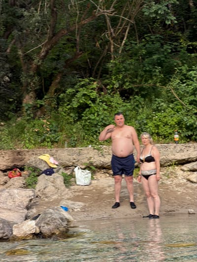 In Spiljani, Bosnia and Herzegovina, on a rocky bank beside a body of water, a man and a woman stand near the water's edge. The man, wearing dark blue swim shorts, has his right arm raised. The woman, in a black bikini, has her left hand near her hip. Behind them, several large rocks are visible, with clothing items and bags scattered on and around them. The background is filled with dense green foliage and trees. A bottle of orange liquid stands on a rock formation in the mid-ground. The water in the foreground is clear and reflects the surrounding environment.