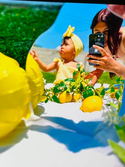 This is a close-up, vertically oriented photograph captured on a sunny day, likely outdoors, given the bright lighting and the presence of greenery. The scene appears to be a reflection, possibly in a screen or polished surface, as the image is mirrored and slightly distorted.
The primary subjects are two individuals. In the foreground, a person's hand is visible, holding a dark smartphone and wearing rings on their fingers. The hand is positioned near a young child who is wearing a yellow outfit and a yellow headband with a bow. The child's face is turned downwards, and they appear to be reaching out. Behind the hand and child, the reflection of another person's head and dark hair is visible.
The setting includes elements of nature. Bright yellow lemons and green leaves, possibly from a lemon tree, are prominently featured, suggesting a Mediterranean or summery theme. Large yellow floral elements or objects are in the lower part of the frame, adding to the vibrant color palette. A bright blue area, likely representing the sky, is visible in the background. Shadows are cast on a white surface, indicating direct sunlight.
The overall mood conveyed is cheerful and vibrant, due to the bright colors and sunny atmosphere. There is no visible text in the image. The reflection format, combined with the close-up perspective, creates an intimate and perhaps artistic portrayal of the subjects and their surroundings.