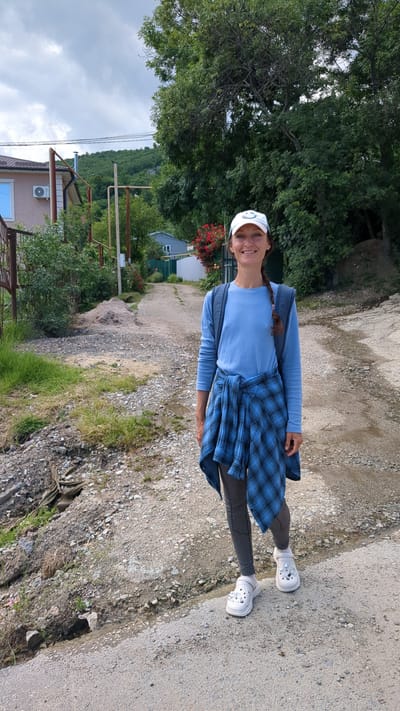 A woman stands on a dirt road in Malyi Mayak, Ukraine. She is wearing a light blue long-sleeved shirt, grey leggings, a blue plaid shirt tied around her waist, and white clogs adorned with decorative charms. A grey backpack is visible on her shoulders. She has a white baseball cap on her head and her brown hair is in a braid. The road is surrounded by vegetation, including trees and bushes, and a residential building is visible in the background. The sky is overcast.