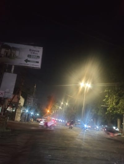 The image captures a street scene at night in Calukan RT 1 RW 5 Keboansikep, Indonesia. A prominent billboard advertises "Breeze Villas" with a price of "590" and a direction arrow indicating "LOKASI" (location). The billboard also shows a phone number. Streetlights illuminate the road, casting bright, starburst-like glows due to the night conditions. Several motorcycles are visible on the road, some with riders, suggesting active movement. One motorcycle in the foreground appears to be pink and white, with a rider wearing a light-colored headscarf. Another motorcycle further down the road is also visible. Trees line the side of the road, and utility poles with wires are present. The overall atmosphere is that of a busy, illuminated street in a residential or commercial area at night.