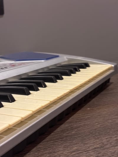 A close-up, low-angle shot shows a portion of a silver electronic keyboard with off-white and black keys. The keyboard is placed on a dark brown wooden surface. The background is a plain, muted gray wall. The image is devoid of any people or specific indicators of location within Novaya Usman, Russia.