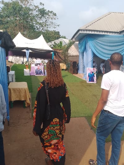 An outdoor event is depicted in Ufuma, Nigeria, under a bright sky with scattered trees in the background. The scene features a central pathway of bare earth flanked by artificial green turf. To the left, a white canopy tent is erected over rows of white plastic chairs arranged on the turf. Black and light blue fabric drapes adorn the tent's interior. A large portrait poster of a man in a white cap and black jacket stands prominently on the turf under this tent, flanked by white floral arrangements. To the right, a building with a silver corrugated metal roof is visible, its front draped in light blue fabric. A large portrait poster of a woman in a blue outfit stands on the turf near this building, with two black loudspeakers adjacent.

In the immediate foreground, facing away from the viewer and walking towards the event setup, is a person with light brown braided hair, wearing a long black garment featuring colorful graphic patterns and text, including "AFRI," "WONDERFUL," and "QUEEN." They carry a small black quilted handbag with a chain strap over their left shoulder. To their right, also walking away from the viewer, is a male figure wearing a white t-shirt, blue jeans, and camouflage-patterned Crocs. Other individuals are partially visible in the mid-ground, under the blue-draped building and near the tents, appearing to be attendees of the gathering. Several small tables, some draped with gold fabric, are positioned throughout the event area.
