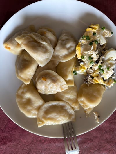 A top-down view displays a white, round plate of food positioned on a textured, dark maroon fabric surface in the city of Des Plaines, United States. The left side of the plate contains approximately 13-15 pale, boiled pierogi, some exhibiting a slightly golden hue, suggestive of caramelized onion or filling elements. The right side features a mound of a salad or side dish, comprising chopped hard-boiled eggs, dark-colored ingredients (consistent with mushrooms), generously coated in a creamy white dressing, and garnished with chopped green scallions. A silver-colored metal fork is partially visible in the lower-right portion of the plate, with its tines resting beneath some of the pierogi.