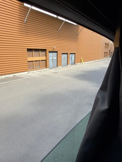The image shows an outdoor scene in Arques, France, featuring a modern building with an orange corrugated metal facade. The building has several doorways, some with gray metal doors. To the left, there is a large window with louvers. The ground in front of the building is paved with asphalt and marked with a white line. A yellow bollard stands near one of the doors. The perspective suggests the photo was taken from inside a vehicle, with a dark fabric curtain obscuring the right side of the frame. The lighting indicates it is daytime.