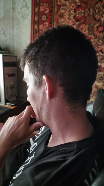 A person is shown from the side, viewed from the back and slightly to the right. They are wearing a black t-shirt with a white graphic design. The person's right hand is resting on their chin, and their head is turned to the left. In the background, a patterned rug is visible on a wall, and to the left, a computer tower with a red light on it is on a desk. The image appears to be taken indoors. The location of this scene in Kibray, Uzbekistan is not indicated in the visual information.