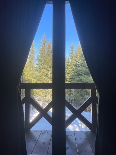The image depicts an outdoor winter scene viewed from an interior perspective, framed by dark curtains partially open on the left and right, and vertical window/door frames in the center. The location is Palež, Montenegro.
The upper half of the view reveals a clear, bright blue sky above a dense forest of evergreen trees, likely conifers, which appear lush and well-lit by direct sunlight. No clouds are visible.
The lower half of the view shows a snow-covered ground extending into the forest. Long shadows are cast across the snow, indicating a clear, sunny day with a relatively low sun angle. In the foreground, a wooden balcony or deck is visible. Its floor consists of dark wooden planks, and it features a wooden railing with a distinctive "X" cross-hatch design, constructed from dark stained timber. Faint strings of what appear to be decorative lights are draped along the railing. No individuals or animals are present in the frame.