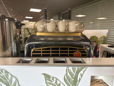 The image displays a coffee machine with a chrome and black exterior, featuring two black handles on top and a gold accent strip. Stacked on a clear glass tray are four pairs of white ceramic cups. Below the cups, a black grille with gold horizontal bars is visible, and a circular logo with a "P" inside it and the word "RESIDENT" is located on the right side of the grille. In front of the coffee machine, a white counter has three square cutouts. One cutout displays the word "FAEMA" in black lettering. The counter is decorated with green leaf patterns. To the left of the coffee machine, a large stainless steel container is present. In the background, through a glass partition, a promotional image of a couple is visible, and behind it, office-like lighting and blinds can be seen. The scene appears to be set within an indoor establishment, possibly a cafe or office break room in Staphorst, Netherlands.