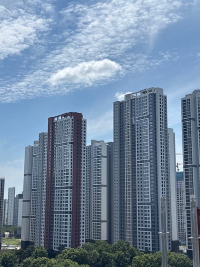 This is a wide shot, taken from a low angle, of a dense urban landscape with numerous high-rise apartment buildings dominating the scene. The buildings are modern and vary in height and design, with predominantly grey and white exteriors, accented by some reddish-brown features on one prominent building in the foreground. Many windows are visible on the facades of the buildings. The sky above is a vibrant blue, populated with scattered white, puffy clouds. In the foreground, lush green trees form a dense canopy, partially obscuring the lower sections of the apartment buildings and suggesting a park or residential area at the base of the towers. A construction crane is visible in the distance, indicating ongoing development in the area. The image appears to be taken during the daytime, with bright sunlight casting shadows. The scene depicts a densely populated residential district within a city.