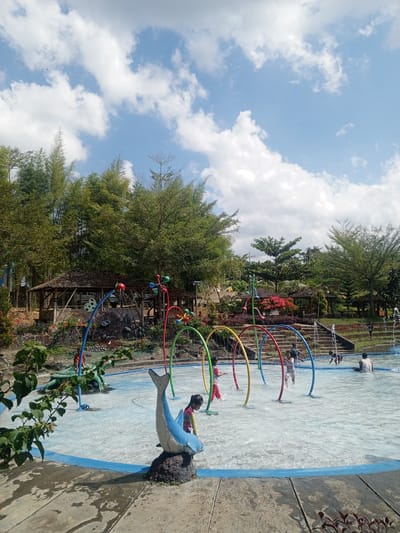 The image depicts a lively water play area in Darsono, Indonesia, under a bright, partly cloudy sky. In the foreground, a shallow pool is filled with clear water, featuring colorful archways that spray water. Children are actively engaged in the water, some running and splashing, while others sit or stand near the spray arches. A blue dolphin statue on a rock is positioned prominently to the left. Beyond the water area, a tiered landscape rises, with various plants and trees, including bamboo, creating a lush backdrop. Traditional Indonesian-style huts with thatched roofs are nestled into the hillside, providing shade. In the distance, more greenery and trees are visible, suggesting a natural, park-like setting. The overall atmosphere is one of recreation and enjoyment on a sunny day.