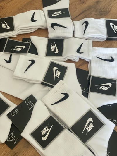 The image shows a pile of white Nike socks with black Nike logos, laid out on a wooden surface. Some of the socks are still in their original packaging with black tags displaying the Nike logo and size information, such as "41-47". The socks are neatly folded and stacked, with some appearing slightly creased. The lighting suggests an indoor environment. There are no people or specific landmarks visible to indicate a location in Krasnyi Krym, Russia.