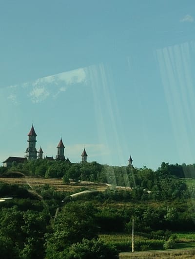 The image shows a castle with multiple spires, set against a clear blue sky. The castle appears to be on a hill, surrounded by trees and greenery. The overall impression is one of a picturesque landscape. The castle is a prominent structure, with its pointed spires and detailed architecture. The surrounding environment is lush and natural, with a sense of tranquility. The image does not contain any people or actions. The specific location in Roșiori, Romania cannot be determined from the image alone.