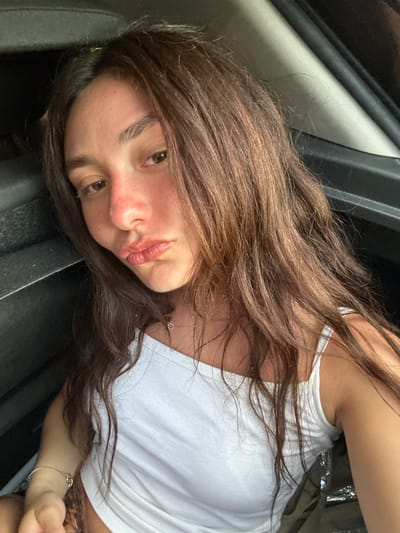 An indoor, close-up, front-facing image features a single individual, a female, seated within the interior of a vehicle. Her long, dark brown hair cascades over her shoulders and chest. Her face exhibits a reddish hue on the nose and cheeks, consistent with a sunburn. Her lips are pursed in a pout, and her gaze is directed towards the camera. She wears a white, thin-strapped tank top, a delicate chain necklace with a small circular pendant, and a light-colored bracelet on her left wrist. A small portion of an animal print garment is visible beneath her white top. The background consists of dark, indistinct elements of the vehicle's interior, including a dark seat or headrest to her left and a car door frame to her right. The specific location within the city of Deià, Spain, is not discernible from the provided visual information.
