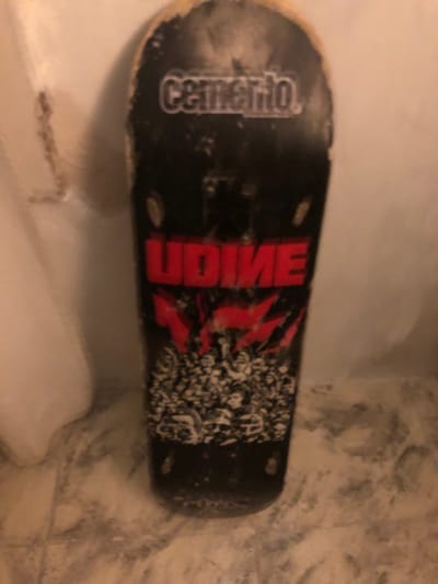 The media file displays a horizontal image of a used skateboard deck, seen from above. The skateboard is resting on a light-colored, possibly tiled or textured floor or surface. The overall lighting suggests an indoor setting, likely under artificial light.
The skateboard deck is predominantly black, covered with grip tape, showing signs of wear, including dust and scuff marks, particularly around its edges.
Visible text on the deck includes:
- On the left side, in white stylized letters, the word "Cemario" or "Cenario" is legible, though slightly blurry.
- In the center, large, bold red letters spell out "UDIME".
- To the right of the "UDIME" text, there is a complex graphic design in black and white/grey, featuring what appears to be abstract patterns or possibly intertwined skull-like imagery.
No people or activities are present in the image; the skateboard is stationary. There are no specific visual cues within the image itself to confirm the provided location context of Beccar, Argentina.