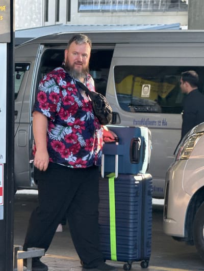 A middle-aged man with a beard wears a floral shirt and black pants. He stands next to stacked luggage with a lime-green strap. A black bag rests on his chest. He faces the camera with a slight smile. A silver van is behind him with Thai writing visible on the window. Another person is partially visible behind the van.