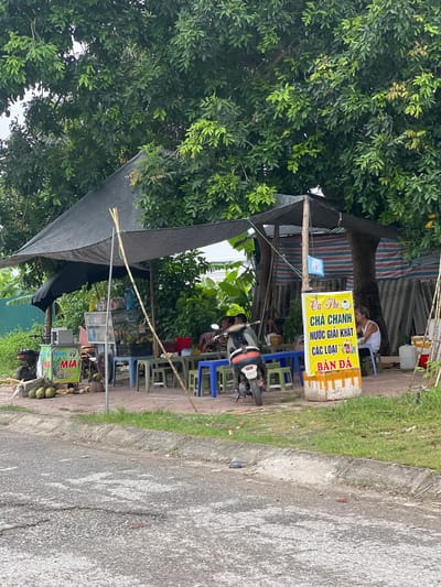 An outdoor refreshment stand is situated next to a road, under the canopy of large trees. The stand features a black tarp roof supported by wooden poles. In front of the stand, several plastic tables and chairs are arranged. A motorcycle is parked to the left of the stand. A yellow sign with Vietnamese text and images of drinks is prominently displayed. Coconuts are visible in a crate near the stand. To the right, a woman is seated at a table, and another person is partially visible. The environment appears to be in Phúc Yên, Vietnam, suggested by the style of the stand and the language on the signage. The ground in the foreground is paved with asphalt, and a grassy median separates it from the stand. The overall scene depicts a typical roadside vendor in a tropical or subtropical urban setting.