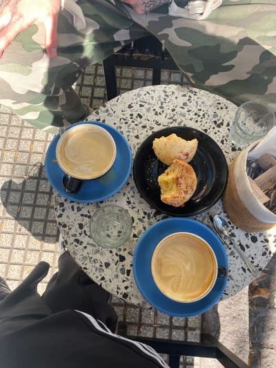 The image depicts a table at a cafe in Olivos, Argentina. The table, which has a light gray speckled surface, holds a black plate with two partially eaten cheese breads, two blue cups of coffee, one empty glass, one glass with remaining water, a small woven basket containing sugar packets, and one spoon. A person wearing camouflage pants is seated on the left, and another person wearing black Adidas pants with white stripes is seated on the right. The table is positioned outdoors on a tiled surface.