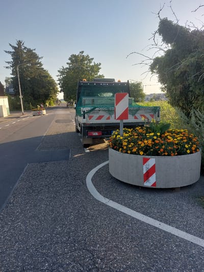 The image shows a street scene in Herzogenrath, Germany. In the foreground, a concrete planter filled with yellow and orange flowers is placed on the asphalt. The planter has red and white reflective markers. Behind the planter is the rear of a white truck covered with a green net. The truck has red and white reflective markers on its tailgate. The street has a white line painted on the asphalt. Trees line the street and the sky is clear and blue. There are houses and buildings visible in the background. The scene appears to be a public street or road.