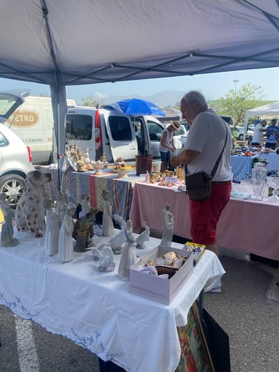 This is a street market scene in San Luis De Sabinillas, Spain, likely a flea market or antique fair, under a white canopy. In the foreground, a white tablecloth with blue embroidered trim covers a table laden with porcelain figurines of people and animals, including a large spotted dog. Behind this table, a man wearing a white t-shirt and red shorts is examining items on a table covered with a patterned cloth. Further back, a white van with "RETRO" prominently displayed on its side is visible, along with other vehicles and market stalls. The background shows a clear sky, distant hills, and more market tents. Several people are browsing at various stalls, suggesting an active market environment.