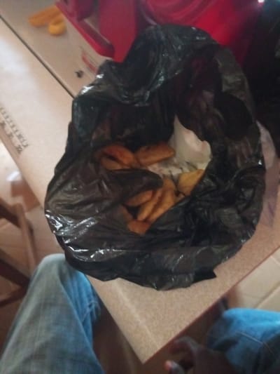 A black plastic bag filled with fried plantains sits on a table. A person wearing blue jeans is partially visible in the foreground. The environment appears to be indoors, possibly a home or a street-side stall in Agbara, Nigeria. The lighting is dim.