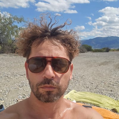 The image is a close-up, selfie-style shot of a man with curly brown hair and a beard, wearing sunglasses with brown frames. He is outdoors on a beach with a pebble and sand surface. In the background, there are trees, a body of water, and mountains under a blue sky with white clouds. The man's torso and shoulders are visible, and his skin appears wet or sweaty. Two towels, one green and one orange, are laid out on the beach behind him. The overall impression is that of a person enjoying a sunny day at the coast. The location is identified as Astros, Greece, suggesting a coastal setting within that region.