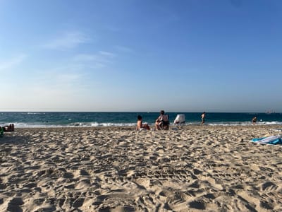 The image depicts a daytime beach scene at Palmachim, Israel. A clear, light blue sky with minimal wispy clouds dominates the upper portion of the frame. The Mediterranean Sea occupies the middle ground, displaying dark blue water with white waves breaking near the sandy shoreline. A large cargo ship or similar vessel is visible on the distant horizon to the right, along with a faint wake from a smaller, faster moving craft further left.
The foreground consists of light-colored, fine-grained sand marked with numerous footprints and slight depressions. Scattered across the sand are various individuals and objects. On the far left, a person is partially visible lying on their stomach. Towards the mid-left, two individuals are present: one person is seated in a dark-colored folding beach chair, facing away from the camera, while another person sits directly on the sand nearby, also facing away. Adjacent to them is a light-colored baby stroller.
Further to the right, closer to the water's edge, a lone individual is walking through the shallow water. To their right, another person is seen playing with a white ball at the water's edge. Within the deeper water, at least two heads are visible, indicating swimmers. On the far right foreground, a deflated or rolled-up blue and white inflatable object lies on the sand. In the far left foreground, a green object, possibly a bag or towel, is partially visible. The overall scene suggests recreational activities on a sunny day.