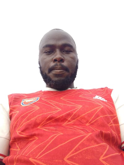 A medium shot captures a dark-skinned man with a shaved head and a full, dark beard. He is wearing a red and white Arsenal soccer jersey with the Adidas logo. The background appears to be a plain white or very light overcast sky. The picture gives the impression of being taken from a lower angle. The location is unspecified within the city of Mulke Barikin Sayejo, Nigeria.