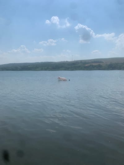 The image depicts a person on a paddleboard in a body of water. The person is positioned in the middle of the frame, standing on the board which is partially submerged. The water is calm with ripples visible on the surface. In the background, a lush green hillside stretches across the scene, its slope covered in trees and vegetation. The sky above is a pale blue, dotted with fluffy white clouds. The overall scene suggests a peaceful, outdoor activity on a clear day. There is no visible text in the image.