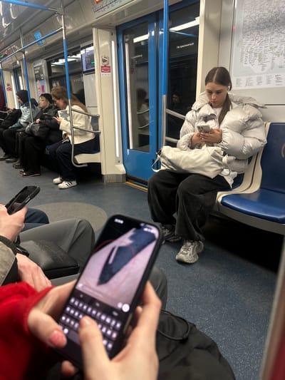 The image shows the interior of a subway car, likely in the city of Dubrovki, Russia. A young woman in a white puffer jacket, black pants, and grey sneakers is seated on a blue subway seat and looking at her smartphone. Other passengers are visible in the background, some seated and some standing. In the foreground, a person is holding a smartphone, with their fingers touching the touchscreen keyboard. A black Hugo Boss bag is near the hand holding the phone.