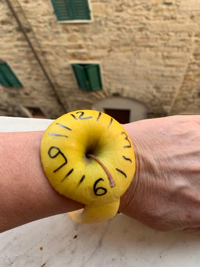 The image shows a close-up of a person's wrist wearing an apple as a bracelet. The apple is yellow and has black markings that resemble clock numbers and hour lines. The markings include numbers like "6", "9", "12", and "21" (possibly representing 9 PM or 21:00 in 24-hour format). There are also several single dash marks indicating hours. The apple's stem is visible.

The background is out of focus but appears to be a stone building with green shutters, suggesting an outdoor setting in a place with traditional architecture. The lighting suggests it is daytime, likely outdoors. The texture of the skin on the wrist is clearly visible, showing wrinkles and pores. The stone wall in the background has a weathered appearance. The overall impression is a playful or artistic modification of an everyday object. The context provided indicates the location is San Quirico d'Orcia, Italy, a town known for its historical architecture.