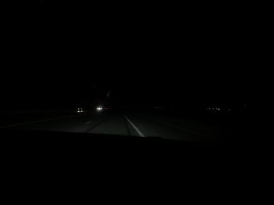 The image shows a night scene under a bridge or overpass, possibly on a highway. There is a faint white line painted on the surface above, indicating a road. Dim lights are visible in the distance, and the headlights of a vehicle are approaching. Due to the limited visibility and lack of specific landmarks, it is impossible to confirm the location as Marmarashen, Armenia, or provide further details about the scene.