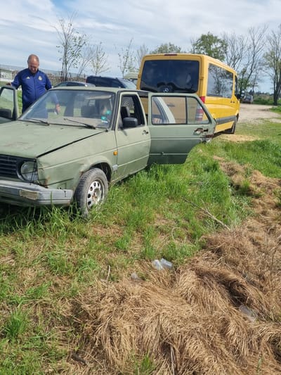 The scene is located in what appears to be a field in Ovcharovo, Bulgaria. An older man wearing a blue jacket is standing near the open door of a dilapidated, olive green car. A yellow Mercedes-Benz Sprinter van is parked nearby. The surrounding terrain consists of grass and dry, matted vegetation. There are also discarded plastic bottles visible on the ground. The condition of the green car suggests it has been abandoned or unused for some time.