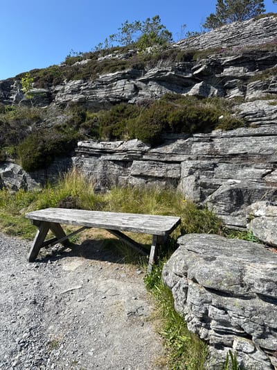 A wooden picnic bench sits on a gravel path next to a rocky hillside covered in heather and sparse trees under a clear blue sky. The bench is weathered and worn, with a simple A-frame design. Large, stratified rock formations dominate the background, suggesting a natural, possibly mountainous or coastal, environment. The scene appears to be a rest stop or viewpoint in the natural landscape of Rong, Norway.