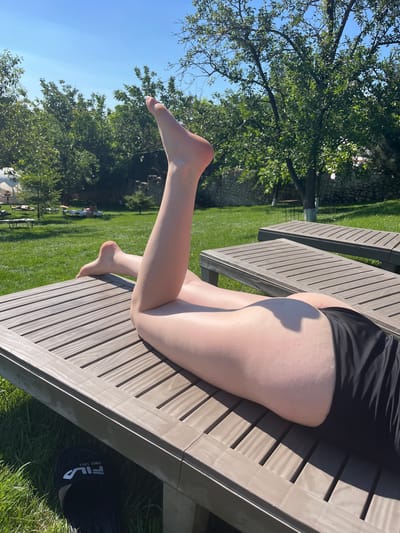 A person is lying on a wooden outdoor bench on a sunny day. The person is wearing black shorts and is positioned with their legs extended. One leg is bent at the knee, with the foot resting on the bench, while the other leg is mostly obscured. The scene is set in a grassy outdoor area with trees and a stone wall visible in the background. The lighting suggests it is daytime, likely late morning or early afternoon, with clear blue skies. A black Fila slide is visible near the bench, with the text "FILA" and "SINCE 1911" printed on it. The overall impression is one of relaxation and enjoying the outdoors.