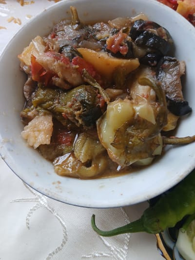 A close-up, overhead shot shows a white bowl filled with a cooked vegetable dish. The dish appears to be a stew or ratatouille, with visible pieces of eggplant, bell peppers, tomatoes, and possibly potatoes. The vegetables are in various shades of green, purple, red, and yellow, and are coated in a savory sauce. The bowl has a thin blue rim and sits on a white tablecloth with an intricate embroidered pattern. To the right of the bowl, a single green chili pepper with a curved stem rests on the tablecloth, partially obscuring a decorative dark bowl with blue and white geometric patterns. The lighting is natural, casting soft shadows and highlighting the textures of the food. This dish is commonly prepared in the city of Gulistan, Uzbekistan.