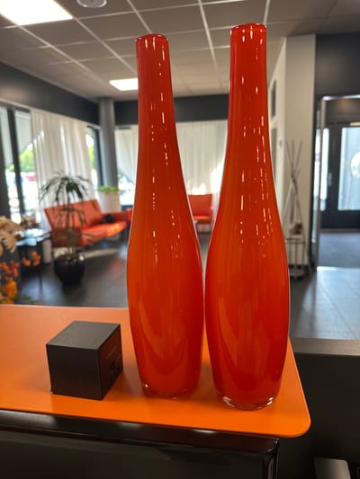 In the city of Staphorst, Netherlands, two tall, vibrant orange glass vases stand prominently on an orange surface. The vases are identical in shape and color, with a glossy finish that reflects the light. To the left of the vases is a small, dark gray, cube-shaped object with visible buttons and ports, suggesting it might be an electronic device. The background is blurred, revealing an interior space with white curtains, a modern orange sofa, and a plant. The ceiling features a grid of recessed lighting. The overall scene is well-lit and appears to be an indoor setting, possibly a showroom or a modern living space.